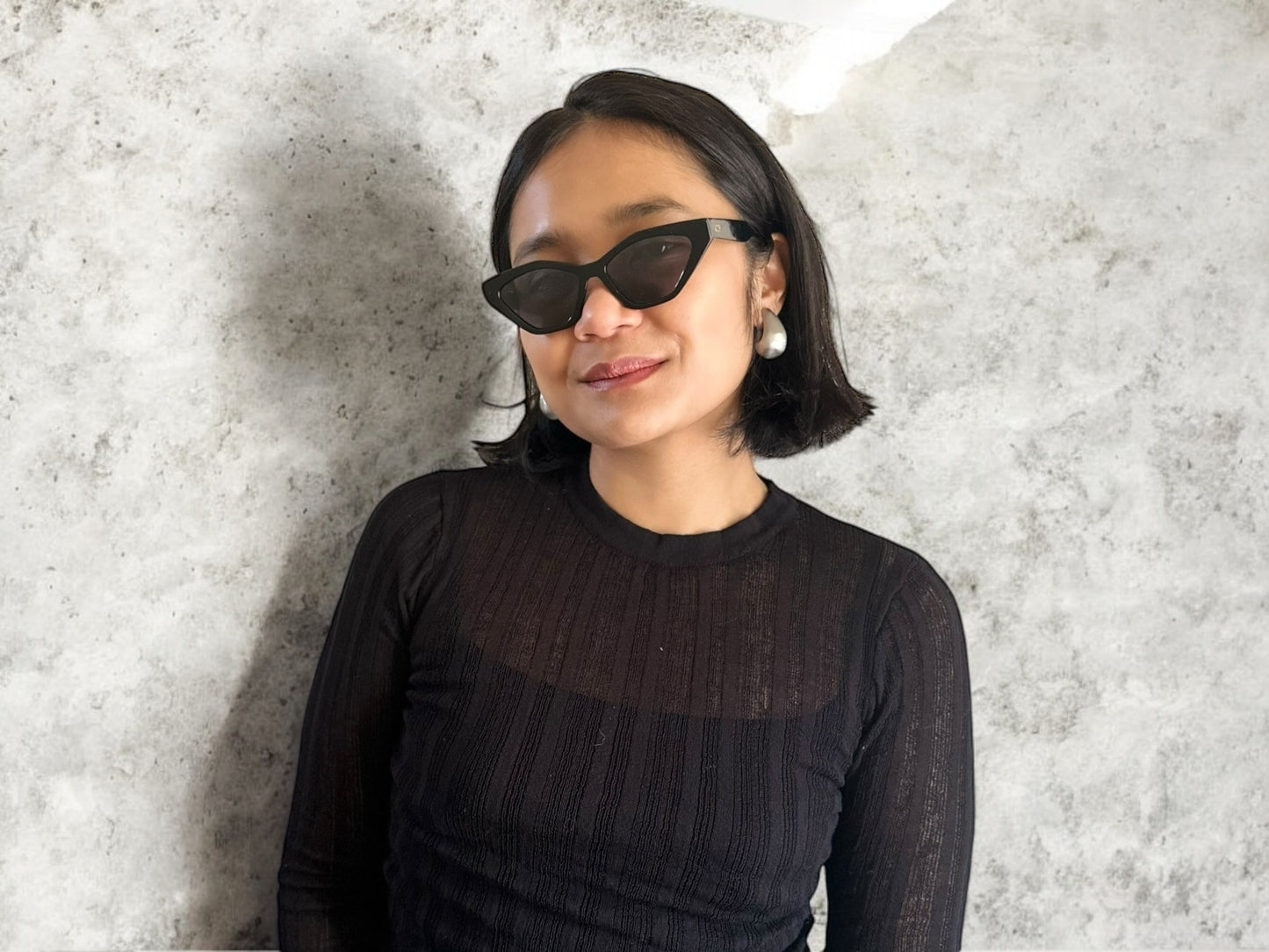 Model wearing Serena Cat Eye Polarised Acetate Sunglasses in Black against a textured stone background, featuring a close-up of the sunglasses and simple, elegant style.