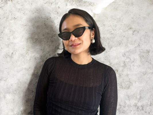Model wearing Serena Cat Eye Polarised Acetate Sunglasses in Black against a textured stone background, featuring a close-up of the sunglasses and simple, elegant style.